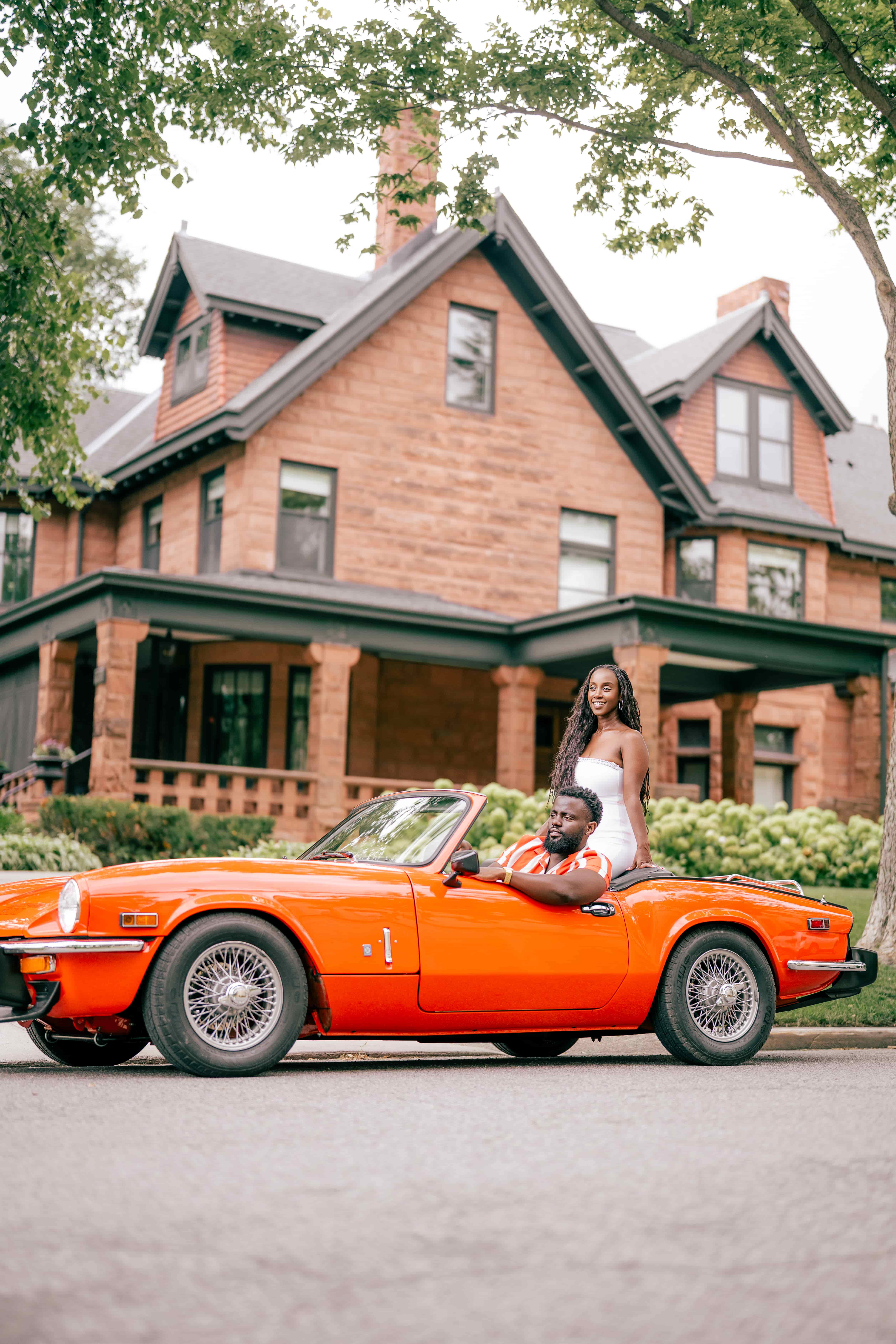 Couple posing with a vintage orange convertible in front of a historic red-brick mansion, editorial couples session
